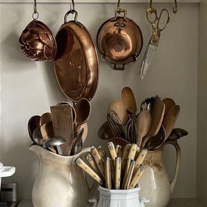 Può includere: Una collezione di utensili da cucina che include stampi in rame, una padella in rame e forbici in metallo appese a una barra in ottone. Due brocche in ceramica color crema contengono cucchiai di legno e fruste, un vaso bianco contiene posate.