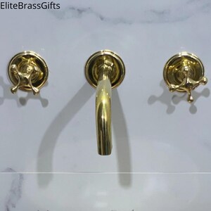 May include: A gold-coloured bathroom tap set featuring a curved spout and two cross-shaped handles. The tap is mounted on a white marble-patterned surface. The brand name "EliteBrassGifts" is visible in the top left corner.