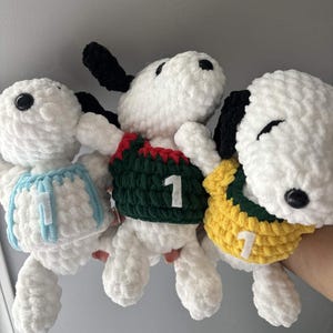 May include: Three white crocheted plush dog toys with black ears and black eyes. Each toy wears a different colored sweater: light blue, dark green with red trim, and yellow with green trim. Each sweater features a white felt number one on the front.
