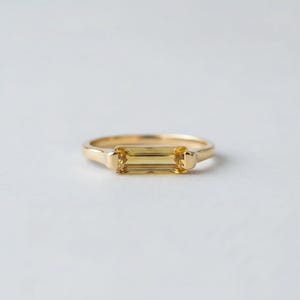 Può includere: Un anello d'oro con una gemma gialla rettangolare. L'anello ha una fascia semplice e un'incastonatura che tiene saldamente la gemma. L'anello è posto su uno sfondo bianco.