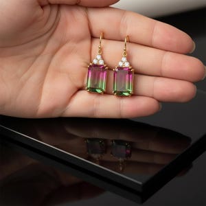 Puede incluir: Un par de pendientes colgantes dorados con piedras preciosas rectangulares bicolor en tonos rosa y verde. Cada pendiente tiene un grupo de pequeñas piedras transparentes en la parte superior.