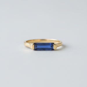 May include: A gold ring with a rectangular, deep blue gemstone. The ring has a simple band with a rectangular setting that holds the gemstone. The gemstone is a vibrant blue colour, and the ring is set against a white background.