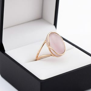 Könnte beinhalten: Ein roségoldener Ring mit einem großen, oval geformten, hellrosa Edelstein. Der Ring ist in einem filigranen, offenen Design gefasst und wird in einer schwarzen Schmuckschatulle mit weißem Innenraum präsentiert. Der Ring ist ein Schmuckstück.