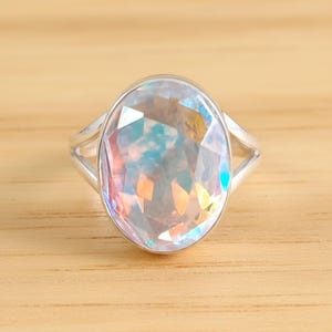 May include: A silver ring featuring a large, oval-cut gemstone with iridescent colors. The gemstone reflects hues of blue, pink, and yellow. The ring's band has a split design on either side of the gemstone.