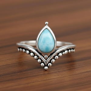 Könnte beinhalten: Ein silberner Ring mit einem tropfenförmigen, hellblauen Edelstein. Der Ring hat ein kronenartiges Design mit kleinen, runden Verzierungen entlang des Bandes. Der Edelstein ist in eine silberne Lünette gefasst.