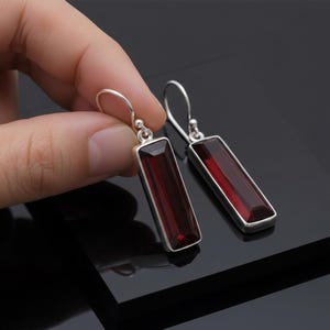 Może przedstawiać: Para srebrnych, prostokątnych kolczyków z głęboko czerwonymi, prostokątnymi kamieniami szlachetnymi. Kolczyki mają prosty, elegancki design z zapięciem na haczyk. Kamienie szlachetne mają bogaty, ciemnoczerwony kolor.