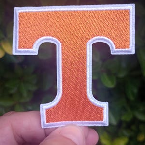 May include: Embroidered orange letter "T" patch with a white border. The patch has a textured appearance and is likely designed to be sewn or ironed onto fabric. The background is a blurred view of green foliage.