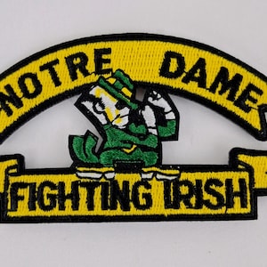 May include: Embroidered patch featuring the Notre Dame Fighting Irish logo. The patch has a yellow banner with the words "NOTRE DAME" and "FIGHTING IRISH" in black lettering. The logo includes a leprechaun in green and white.