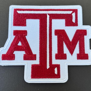 May include: Embroidered patch featuring the letters "A", "T", and "M" in maroon against a white background. The patch has a white border and a rectangular shape with rounded corners. The letters are in a bold, block style.