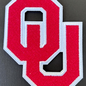 May include: A red and white embroidered patch featuring the letters "OU". The letters are in a bold, block style with a white border. The patch is likely for a sports team or university.