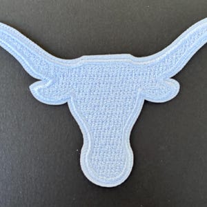 Puede incluir: Parche bordado azul claro con forma de cabeza de toro de cuernos largos. El parche tiene un aspecto texturizado y un contorno blanco. Los cuernos se curvan hacia arriba y hacia afuera. El parche es probablemente para ropa o accesorios.