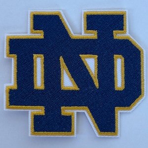 Peut inclure: Écusson brodé avec les lettres "ND" en bleu marine, bordées d'or. L'écusson est posé sur un fond blanc. Il est probablement destiné aux vêtements ou accessoires, représentant une équipe ou une organisation.