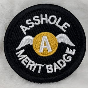 May include: Black circular embroidered patch with the words "ASS HOLE MERIT BADGE" in white lettering. A gold circle with a white "A" and silver wings is in the center.