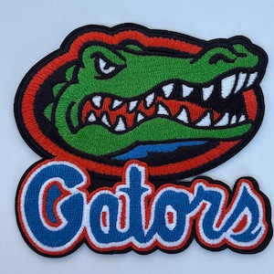 May include: Embroidered patch featuring a green alligator head with white teeth, red accents, and the word "Gators" in blue and white. The patch has a black border with red and orange trim, suitable for apparel or accessories.