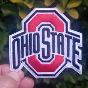 Puede incluir: Parche bordado con las palabras "OHIO STATE" en letras negras sobre una "O" roja. El parche tiene un borde blanco y tiene forma hexagonal. El parche se sostiene frente a un fondo de hojas verdes.