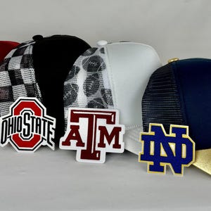 May include: A collection of trucker hats with mesh backs and various team logos. The hats feature a range of colors, including red, black, white, and navy blue, with gold and white brims. Each hat displays a different college team logo.