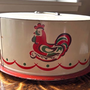 May include: A cylindrical metal cake carrier with a cream-colored body and a red base. The side features a stylized rooster graphic in red, green, and dark brown, with a red scalloped border and green dots along the bottom edge and a metal handle.
