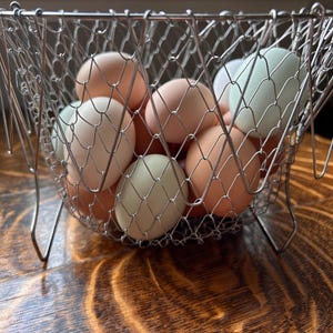 May include: A wire mesh basket filled with a variety of fresh eggs. The eggs are in shades of white, light green, and peach. The basket is made of silver-colored metal and sits on a wooden surface.