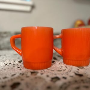 Puede incluir: Dos tazas de vidrio naranja con asas. Las tazas son de un color naranja vibrante y tienen un diseño simple y clásico. Las tazas están sobre un tapete de encaje blanco.