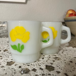 Op de afbeelding: Twee witte keramische mokken met gele bloemmotieven en groene bladeren. De mokken hebben vierkante handvatten en zijn naast elkaar gestapeld. De mokken staan op een kanten kleedje.
