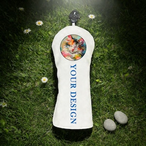 May include: White golf club head cover featuring a colourful cat portrait in a circular frame. The cover has the text "YOUR DESIGN" in blue. The cover is on green grass with small white flowers and two small grey stones.
