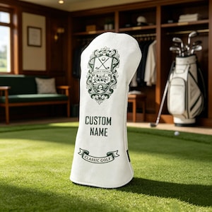 Könnte beinhalten: Eine weiße Golfschlägerhaube mit einem dunkelgrünen Wappen, gekreuzten Golfschlägern und floralen Details. Der Text CUSTOM NAME ist in dunkelgrünen Blockbuchstaben über einem Banner mit der Aufschrift CLASSIC GOLF in dunkelgrüner Tinte gedruckt.