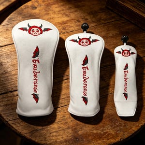 Puede incluir: Tres fundas blancas para la cabeza del palo de golf con un diseño de cara de diablo rojo y la palabra "Exuberance" en escritura roja. Cada funda tiene un tamaño diferente y una parte superior negra.