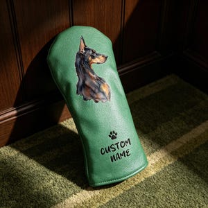 Könnte beinhalten: Ein grüner Golfschlägerhaube mit einer detaillierten Illustration eines schwarz-braunen Dobermanns. Die Hülle zeigt ein kleines schwarzes Pfotenabdruck-Symbol über dem schwarzen Schriftzug CUSTOM NAME, der auf der Vorderseite aufgedruckt ist.