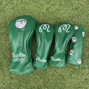May include: Four green golf club head covers with white embroidered details. The covers display the words "Malbon Golf" and a cartoon golf ball character. The covers are arranged on green grass, providing a contrasting background.