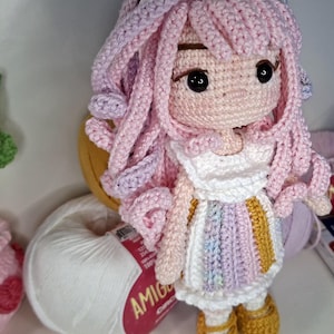 May include: A handmade crochet doll featuring long, curly pastel pink and purple hair, a white unicorn headband with a gold horn, and a dress with vertical stripes in white, pink, purple, and gold. The doll has black eyes and gold shoes.