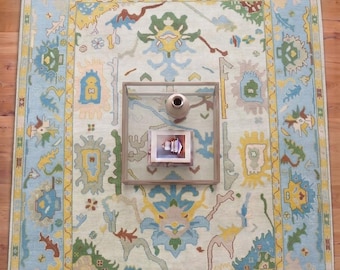 May include: A large, rectangular area rug with a light blue field and a detailed border. The rug features a central design with floral and geometric patterns in yellow, green, and blue. A glass coffee table sits on the rug.