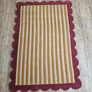 May include: A rectangular rug with a scalloped burgundy border. The rug's interior features vertical stripes in alternating shades of gold and white. The rug is designed for indoor use and adds a decorative element to any room.