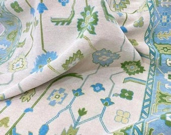 May include: Close-up of a cream-colored fabric with a detailed floral pattern. The design features blue and green floral motifs and geometric shapes. The border is a light blue with a repeating geometric pattern. The fabric is folded, showing the texture and design.