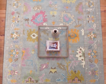 May include: A large, rectangular area rug with a light blue background and a colorful floral pattern. The rug features a border with repeating floral motifs in shades of pink, yellow, and blue. A glass coffee table and vase are on top of the rug.
