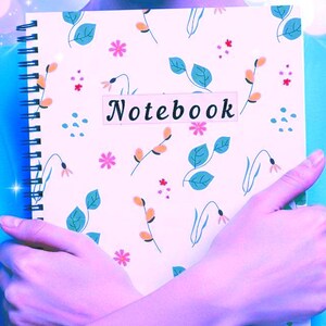 Peut inclure: Cahier à spirales avec un motif floral. La couverture présente un fond rose clair avec des illustrations de feuilles, de fleurs et de brins en orange, bleu et rose. Le mot "Notebook" est imprimé sur une étiquette rectangulaire.