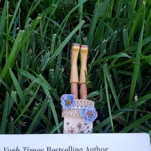 May include: A handmade bookmark with a pair of brown doll legs, a brown and white lace trim, and blue flowers. The bookmark is placed in a book with the text "York Times Bestselling Author" visible.
