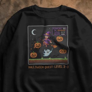 以下が含まれることがあります： ハロウィーンをテーマにしたピクセル化されたグラフィックが特徴の黒いスウェットシャツ。 デザインには、ほうきに乗った魔女、カボチャ、幽霊、そして「HALLOWEEN QUEST: LEVEL 2-3」というテキストが含まれています。 グラフィックはレトロなビデオゲームの美学を持っています。