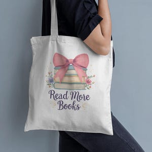 Pode incluir: Saco tote branco com um design que apresenta uma pilha de livros, um laço rosa e elementos florais. O texto "Read More Books" está impresso abaixo. O saco tem um design simples com uma alça de ombro.