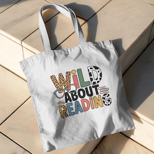 Pode incluir: Saco tote branco com o texto "WILD ABOUT READING" em letras coloridas e estampadas. O saco apresenta um "W" com estampa de leopardo, um "I" verde e um "D" com estampa de vaca em preto e branco. Possui duas alças brancas.