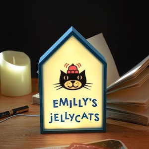 May include: A blue house-shaped night light with the text "EMILLY'S JELLYCATS" below a cartoon cat wearing a hat. The light is on, illuminating the cat graphic and text. A candle and book are in the background.