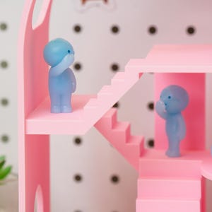 May include: A pink dollhouse with stairs and a balcony, featuring two small blue figurines. The dollhouse has a white background with black dots. The figurines are in a thoughtful pose, with one hand raised to their face.