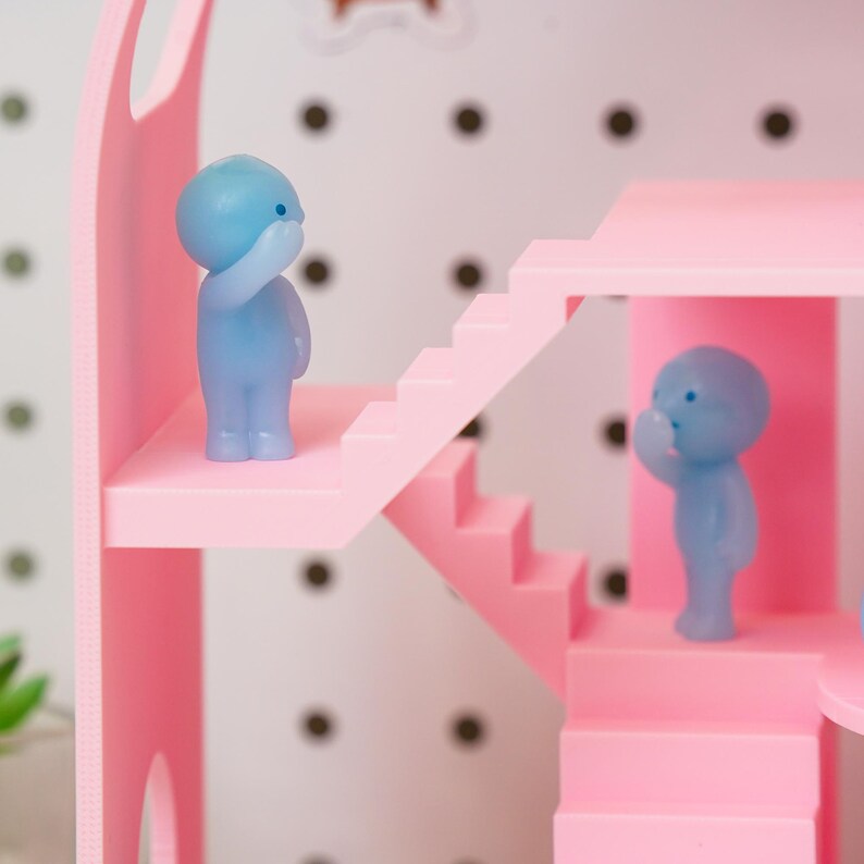 May include: A pink dollhouse with stairs and a balcony, featuring two small blue figurines. The dollhouse has a white background with black dots. The figurines are in a thoughtful pose, with one hand raised to their face.