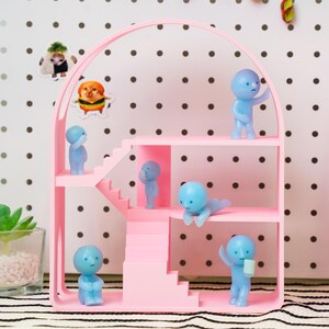 May include: A pink dollhouse-style shelf with multiple levels and stairs, housing several small, light blue figurines in various poses. The shelf has an arched top and is set against a dotted background. Stickers of animals are attached to the background.