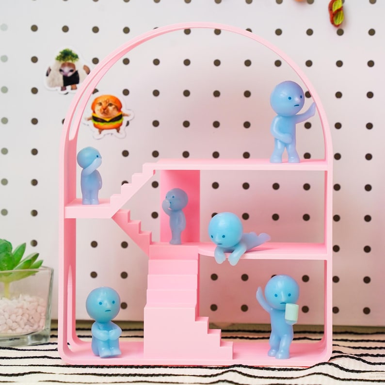 May include: A pink dollhouse-style shelf with multiple levels and stairs, housing several small, light blue figurines in various poses. The shelf has an arched top and is set against a dotted background. Stickers of animals are attached to the background.