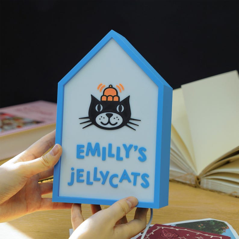 May include: A blue and white house-shaped night light with the text "EMILLY'S JELLYCATS" and a black cat graphic. The cat has a small orange hat and antenna-like lines above its head. The night light is held by a person.