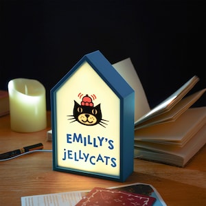 May include: A blue house-shaped night light with the text "EMILLY'S JELLYCATS" and a cartoon cat illustration. The light is on, illuminating the text and cat design. A candle, pen, and open book are in the background.