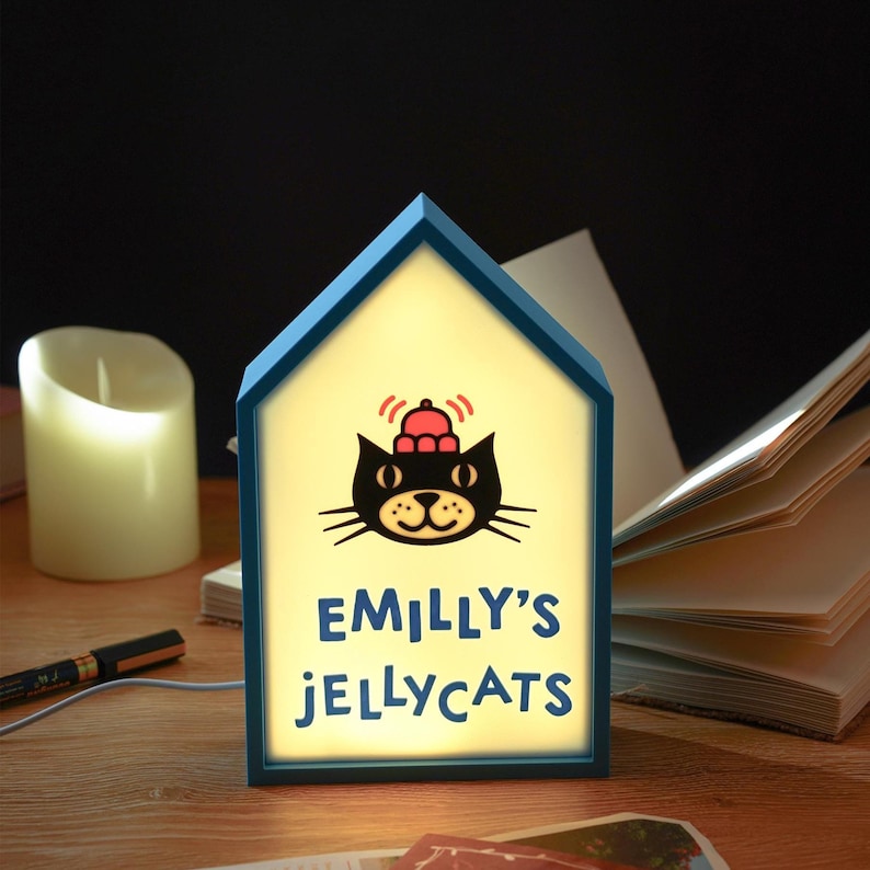 May include: A blue house-shaped night light with the text "EMILLY'S JELLYCATS" below a cartoon cat wearing a hat. The light is on, illuminating the cat graphic and text. A candle and book are in the background.