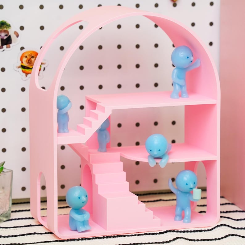May include: A pink dollhouse-style shelf unit with multiple levels and staircases. Several small blue figurines are positioned on the shelves, some standing, some sitting, and one holding a small cup. The unit has an arched top and is made of a solid, light pink material.