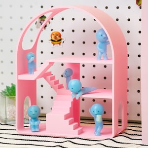 May include: A pink, three-story dollhouse-style shelf with an arched top. The shelf features stairs and multiple levels, populated with small blue figurines in various poses. Stickers of a dog and a cat are on the wall behind the shelf.