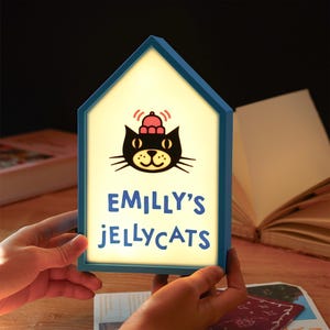 May include: A blue house-shaped night light with the text "EMILLY'S JELLYCATS" and a cartoon cat illustration. The cat is black with a red hat. The light is held by two hands.